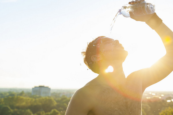 Oberkörperfreier Mann gießt sich Wasser aus einer Flasche ins Gesicht, Sonnenlicht im Hintergrund