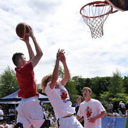 Ein Spieler wirft den Ball in einen Basketball-Korb. Zwei weitere Spieler versuchen das zu verhindern. 